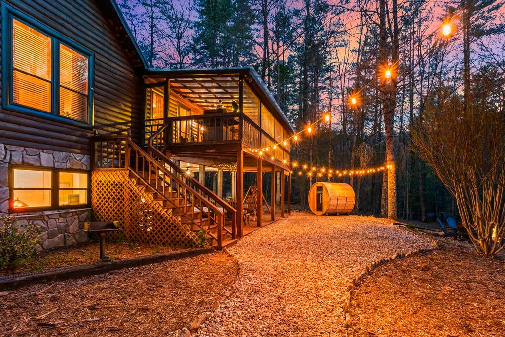 Log cabin with illuminated deck, string lights, gravel path, and barrel structure in a wooded yard at dusk.