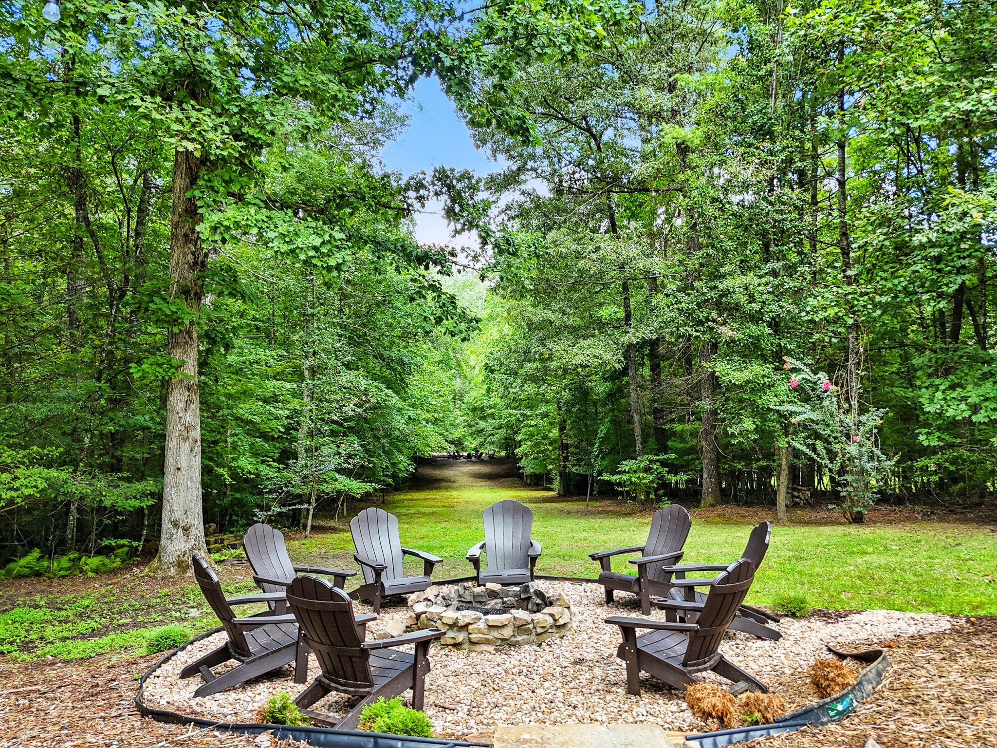 Adirondack chairs circle a stone fire pit in a green wooded backyard clearing.