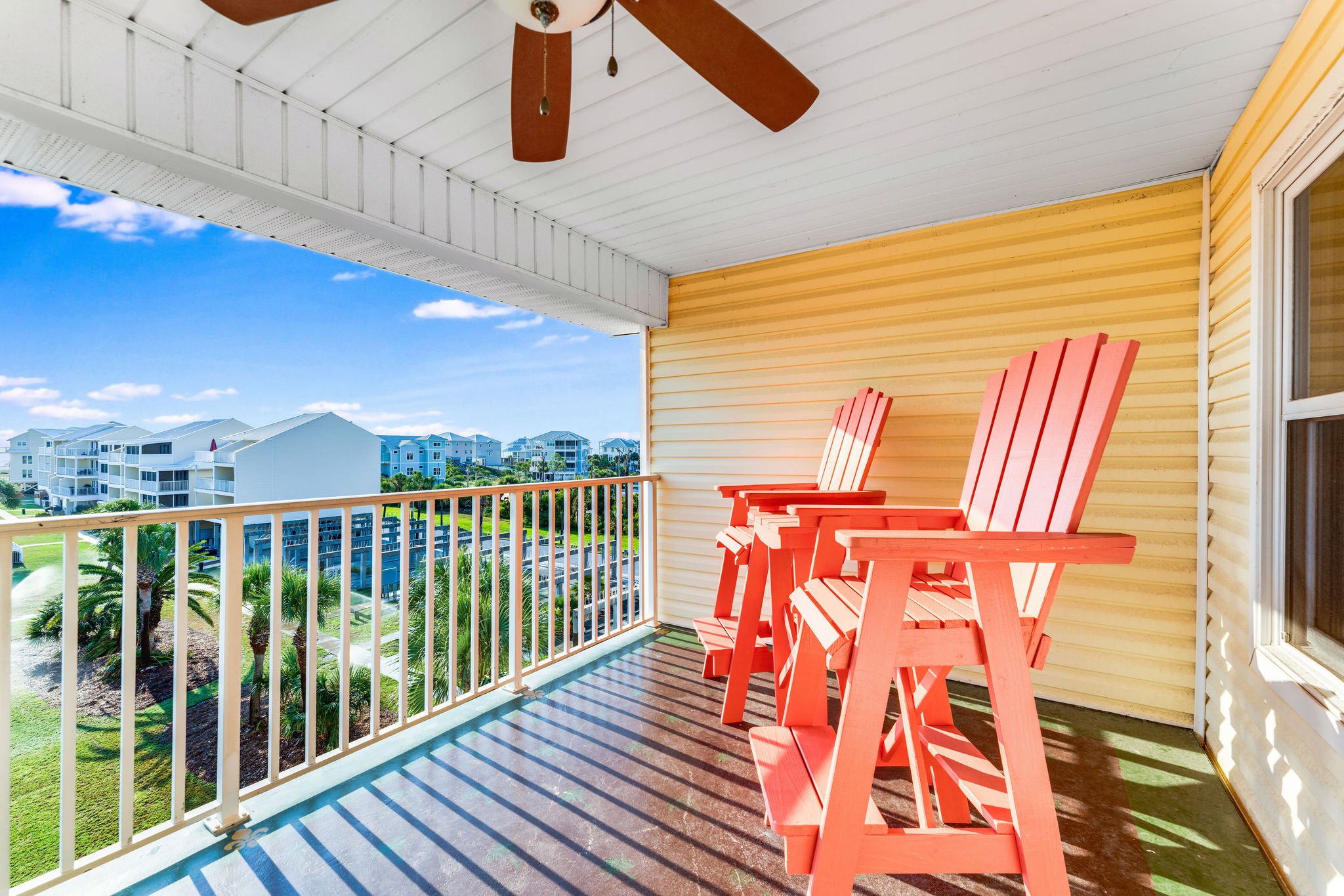 Three coral Adirondack chairs sit on a sunny covered balcony overlooking palm trees and coastal-style buildings.