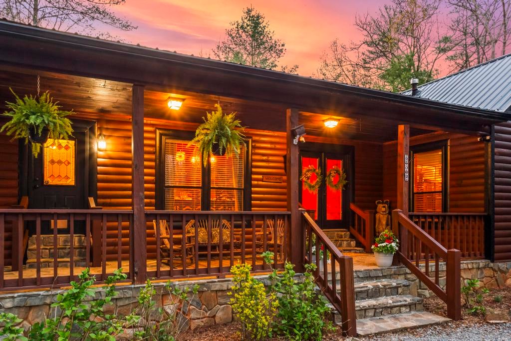 Rustic log cabin porch glowing at dusk, with ferns, wreaths, stone steps, and a pink-orange sky.