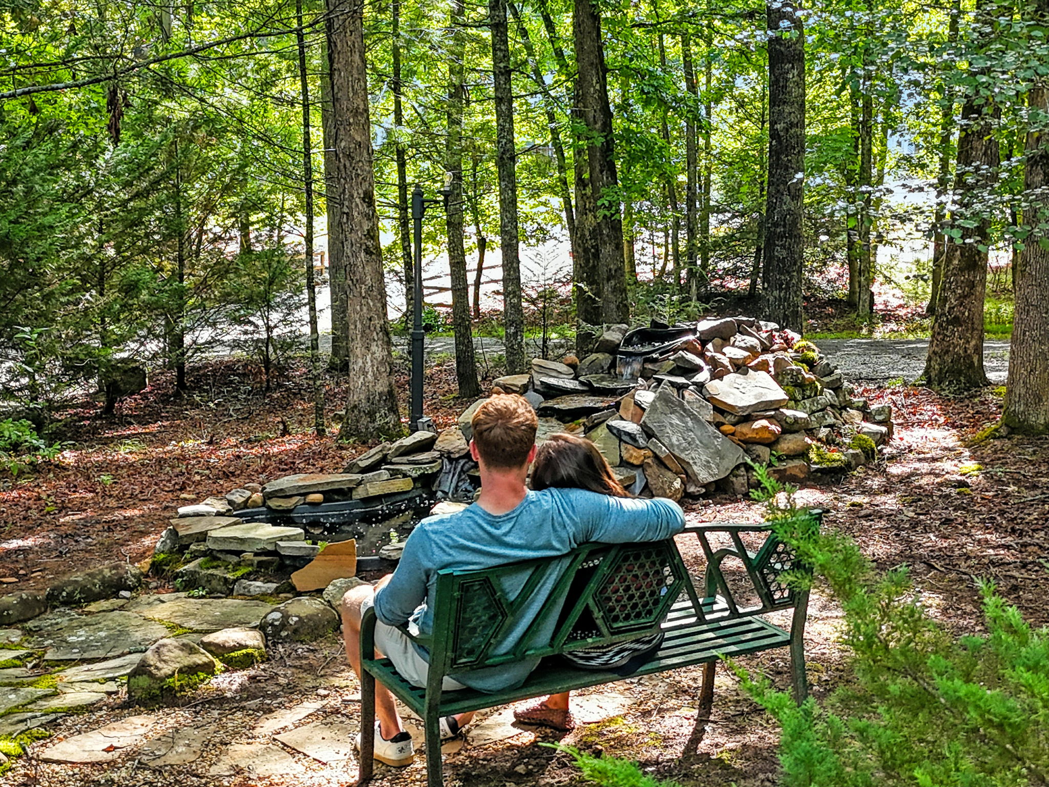 Two people sit on a green bench beside a rock-lined waterfall in a sunlit forest garden.