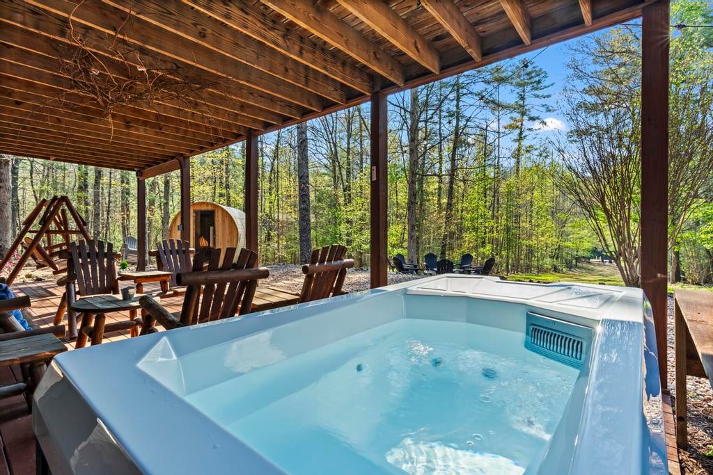 Covered wooden deck with a bubbling hot tub, outdoor chairs, a small sauna, and leafy woodland surroundings.