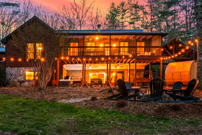 Rustic two-story log cabin glowing with string lights at dusk, with chairs and a barrel sauna in the yard.