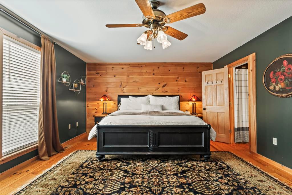 Rustic bedroom with a black bed, wood-paneled accent wall, ceiling fan, patterned rug, and open door.