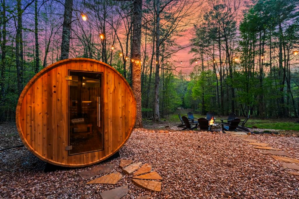 Barrel-shaped wooden sauna, string lights, and a fire pit with chairs in a forest at sunset.