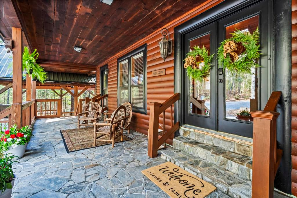 Rustic cabin porch with rocking chairs, fern wreaths, stone steps, and a welcome mat.