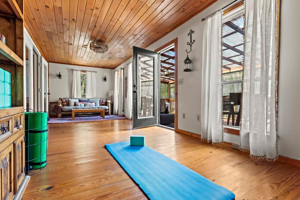 Rustic wood-floored room with a blue exercise mat, sofa seating, open patio door, and sheer-curtained windows.