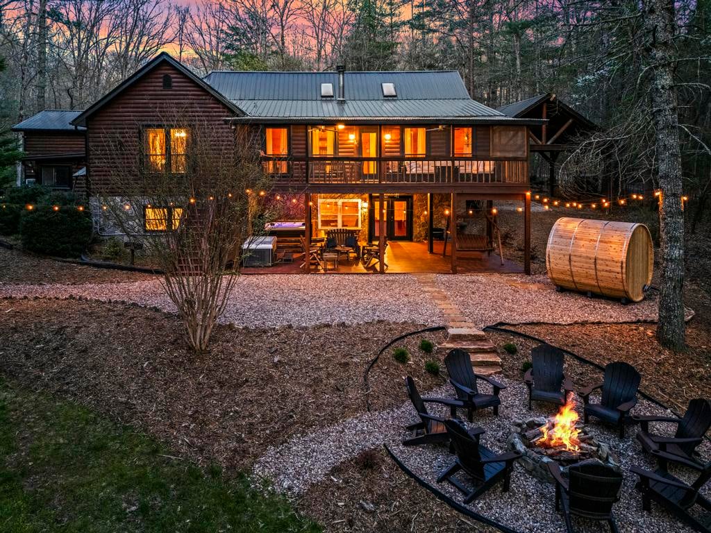 Lit log cabin with string lights, barrel sauna, and fire pit surrounded by chairs at dusk.