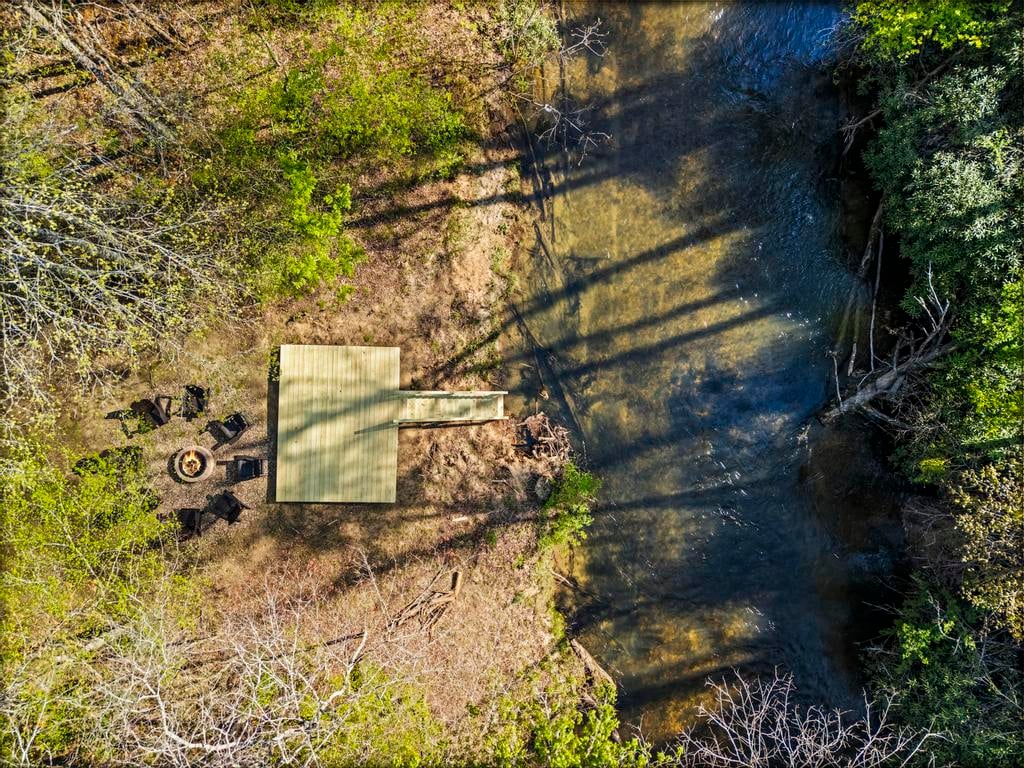 Aerial view of a wooden creekside deck, fire pit, chairs, and shallow wooded stream.