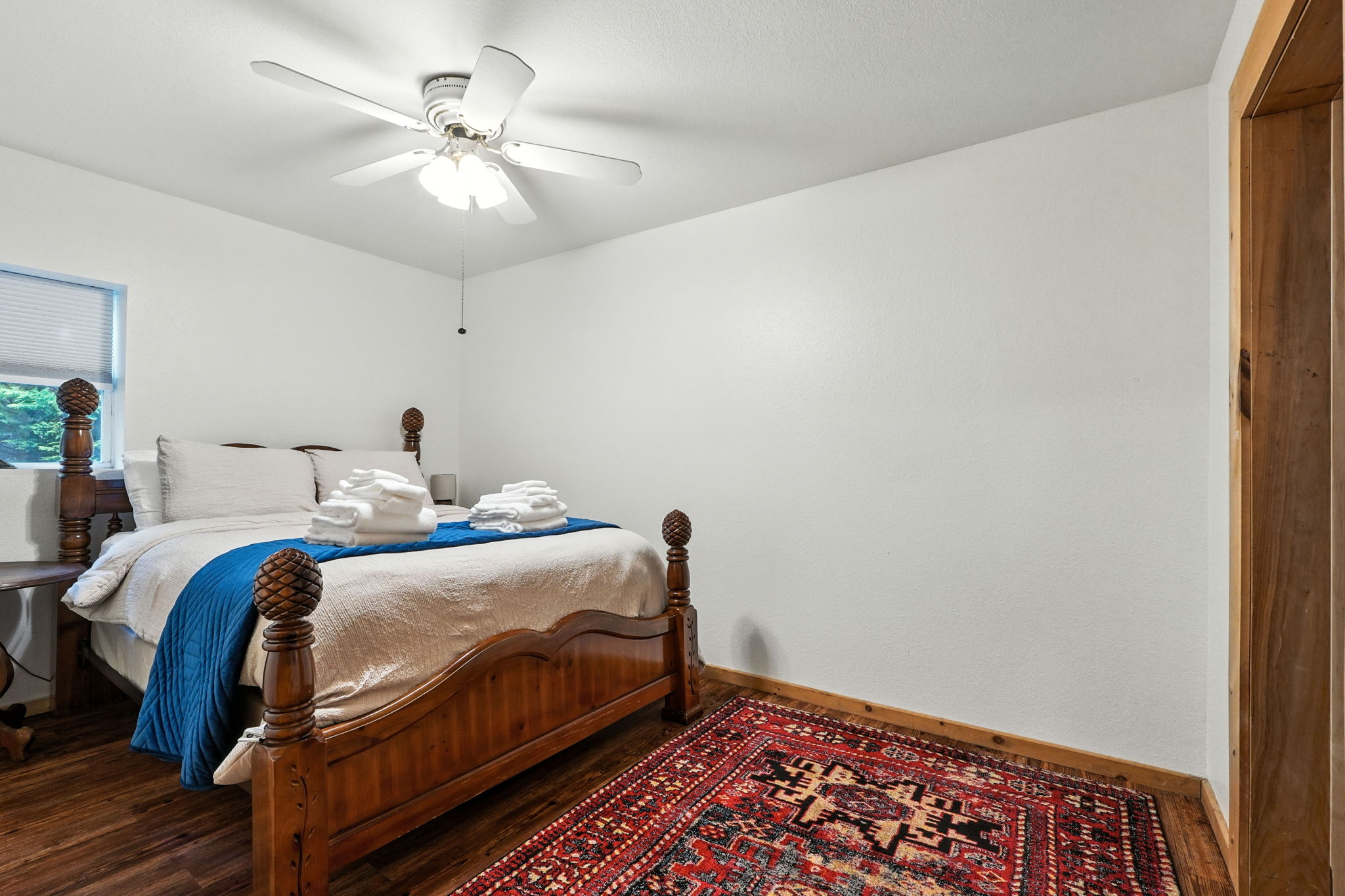 Bright bedroom with a wooden four-poster bed, blue runner, folded towels, ceiling fan, window, and patterned rug.