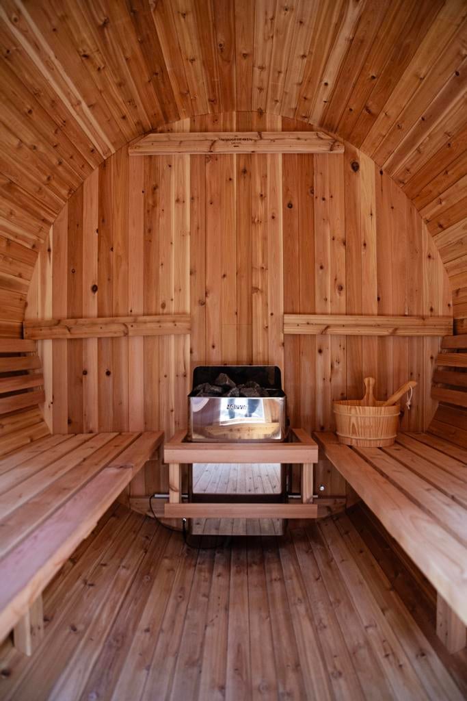 Interior of a barrel-shaped wooden sauna with benches, a central stone heater, and a bucket with ladle.