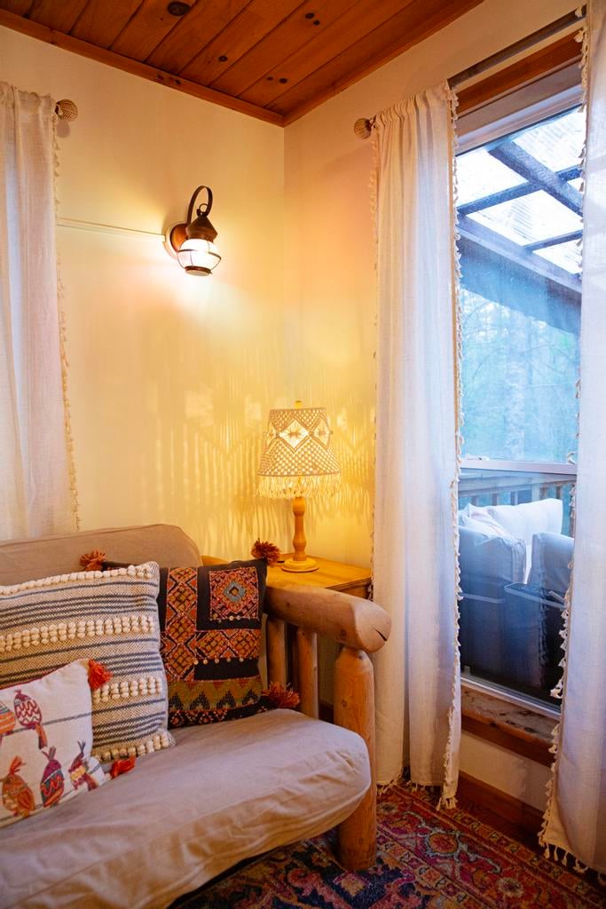 Cozy wood-paneled sitting area with a cushioned armchair, patterned pillows, lamps, rug, and curtained window.
