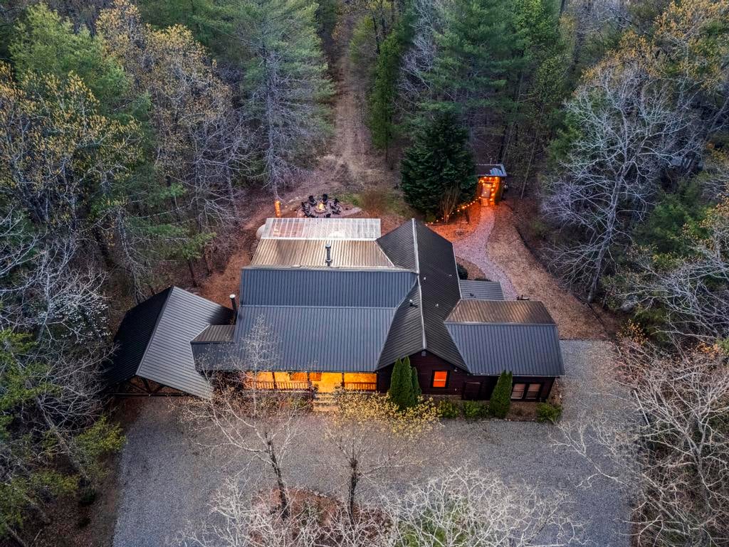Aerial view of a warmly lit dark-roofed cabin in a dense forest at dusk.
