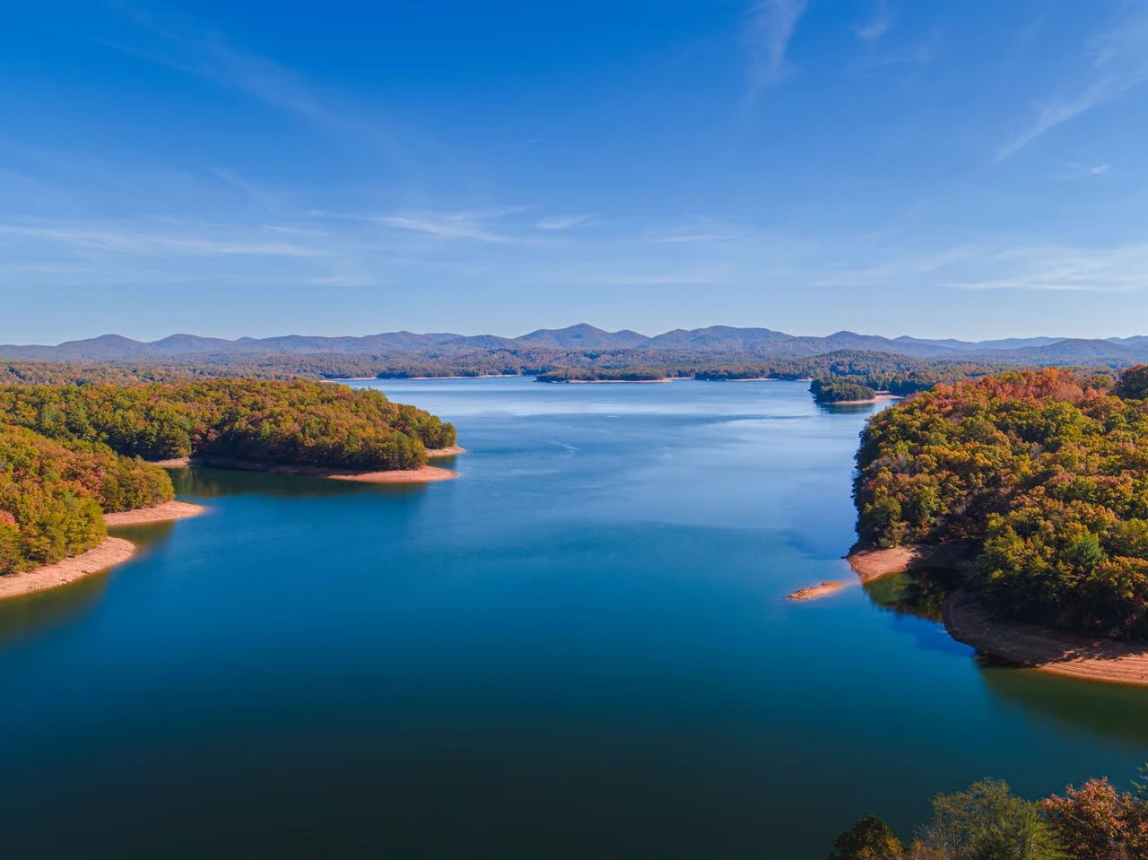 A blue reservoir winding through autumn-colored forest with low mountains beneath a clear, wispy-clouded sky.