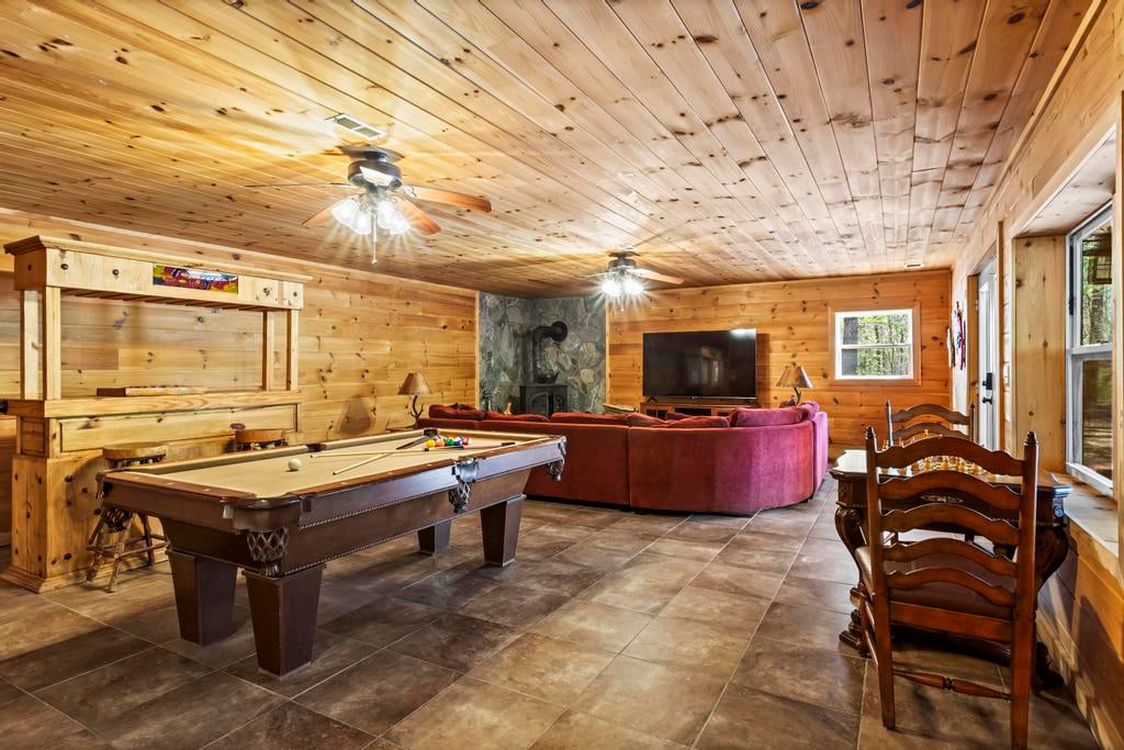 Rustic wood-paneled game room with a pool table, red sectional, fireplace, television, dining chairs, and windows.