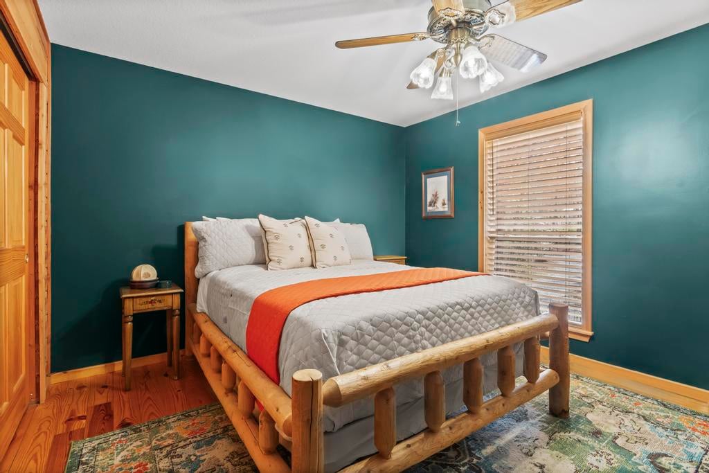 Rustic bedroom with a log bed, teal walls, orange bed runner, window blinds, ceiling fan, and patterned rug.
