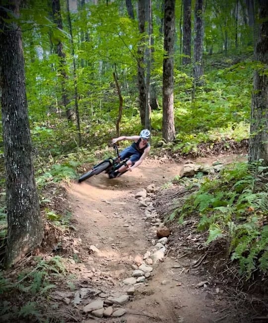 Cyclist leaning through a rocky dirt turn on a mountain-bike trail in a dense green forest.