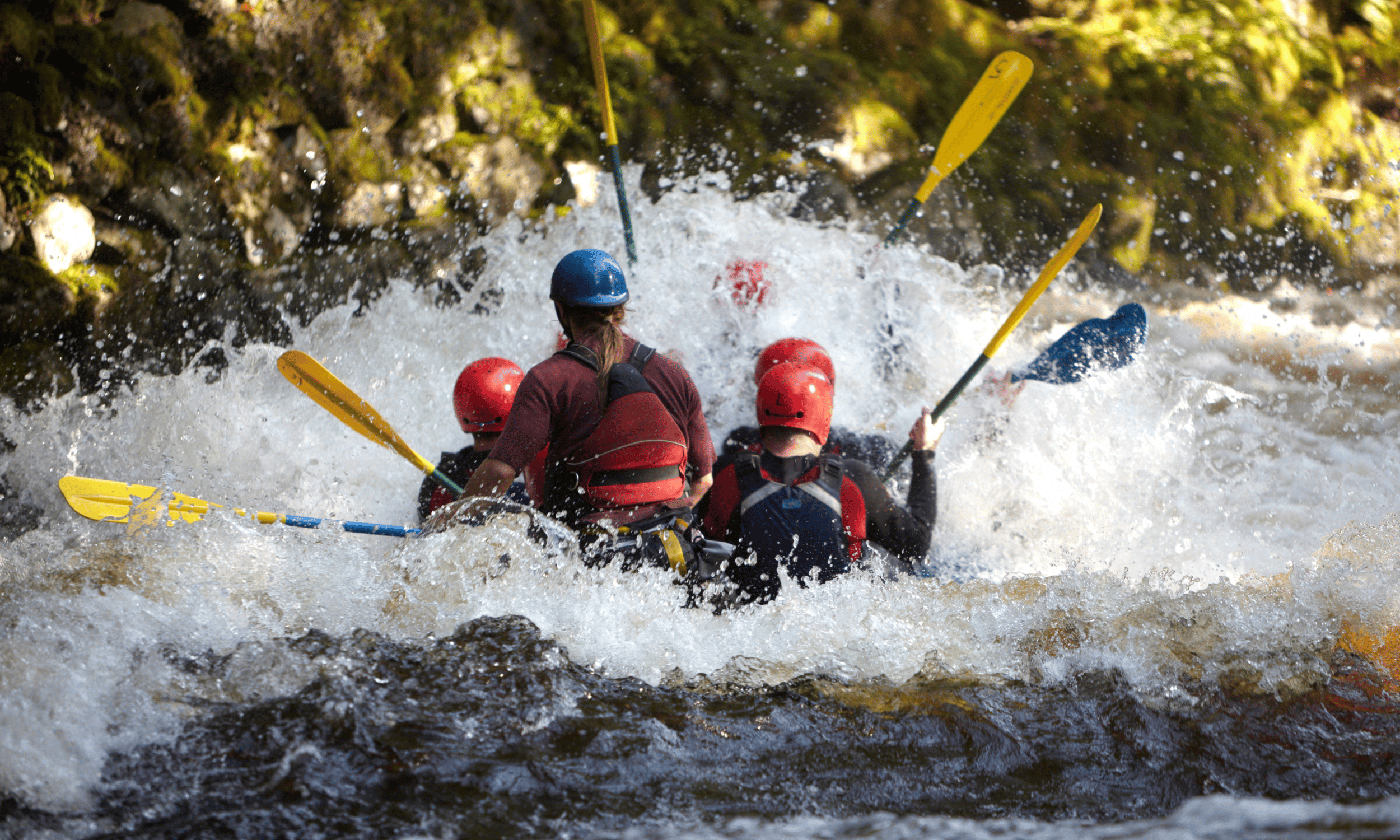 Three helmeted rafters paddle through foaming whitewater among rocky, moss-covered banks.