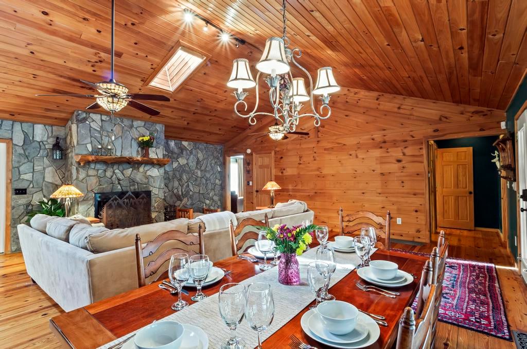 Rustic wood-paneled dining room with a set table, stone fireplace, sofas, chandeliers, and a patterned hallway rug.