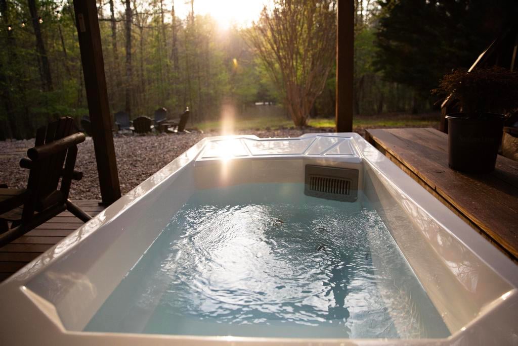 White outdoor hot tub with rippling water on a deck overlooking a sunlit wooded clearing.