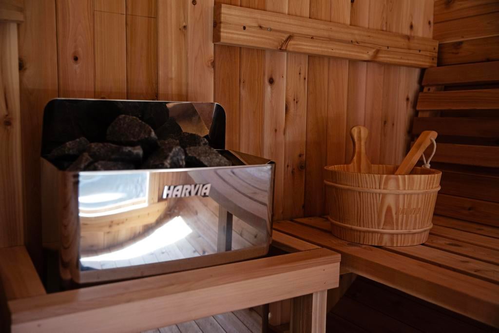 Wooden sauna interior with a stone-filled HARVIA heater, benches, and a wooden bucket with ladle.
