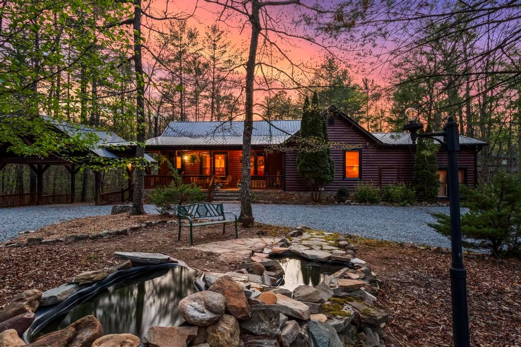 Rustic log cabin glowing at sunset, surrounded by trees, a gravel courtyard, bench, lamp post, and rock-lined pond.