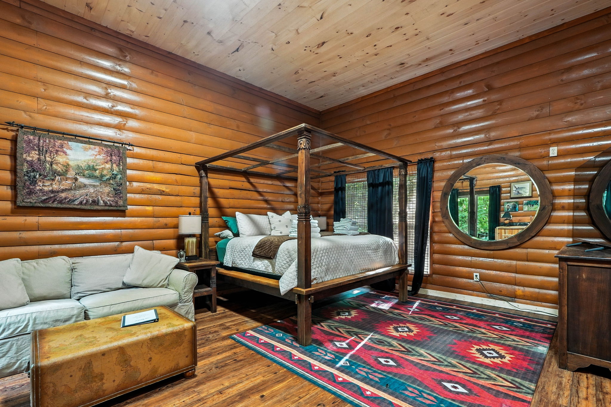 Rustic log-cabin bedroom with a canopy bed, colorful geometric rug, sofa, deer tapestry, and round wall mirror.