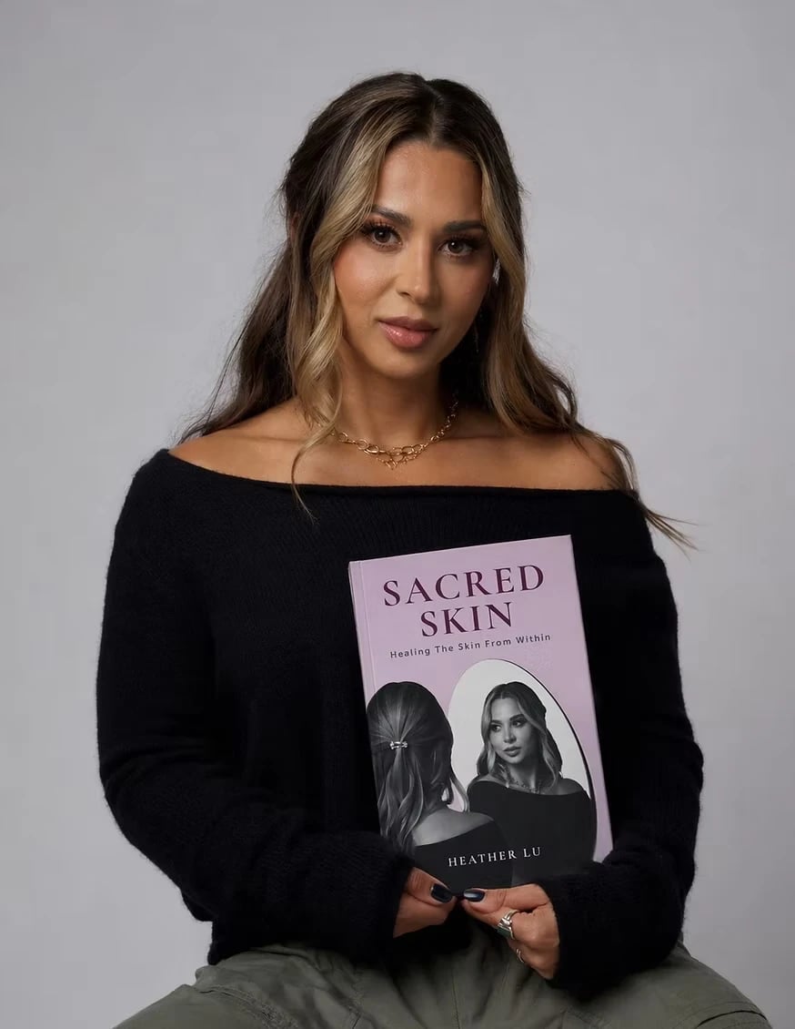 Woman in a black off-shoulder sweater holds a pink book titled “SACRED SKIN” against a gray background.