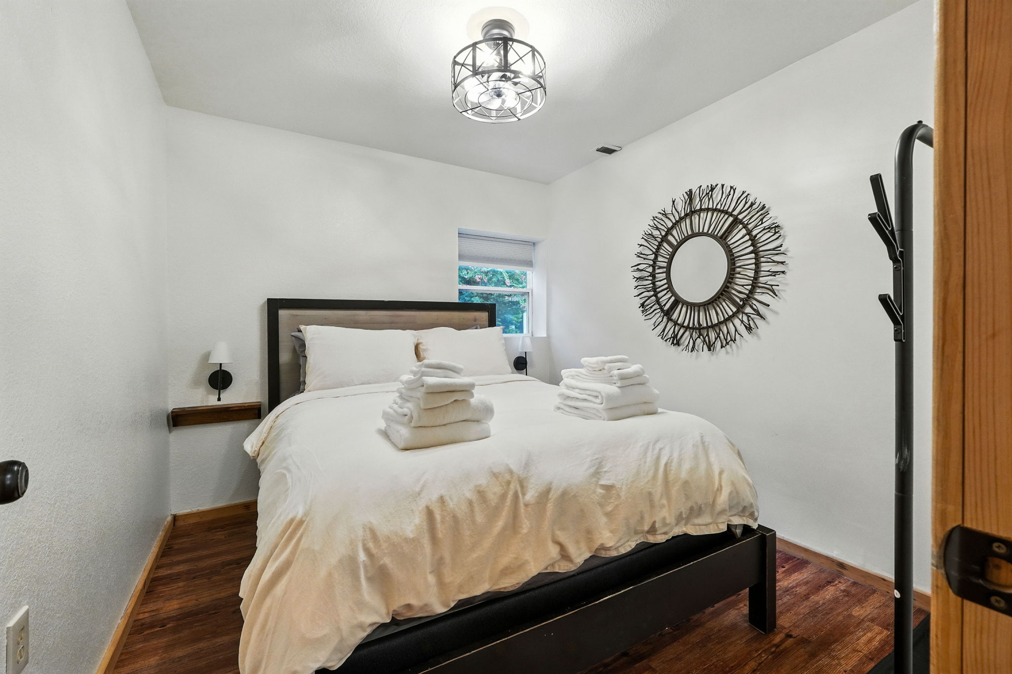 Bright bedroom with a made bed, folded white towels, sunburst mirror, window, and wood flooring.
