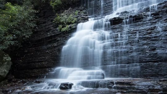 Multi-tiered waterfall flowing over dark layered rocks amid dense green forest.